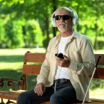 Blind old man wearing earphones listening audiobook, voice message in cellphone