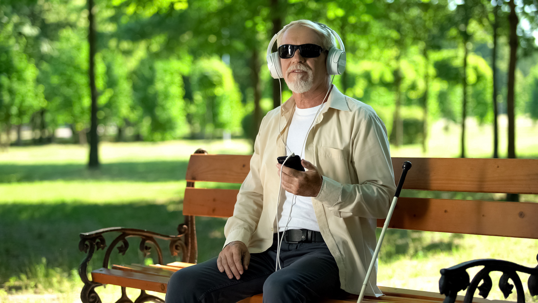 Blind old man wearing earphones listening audiobook, voice message in cellphone